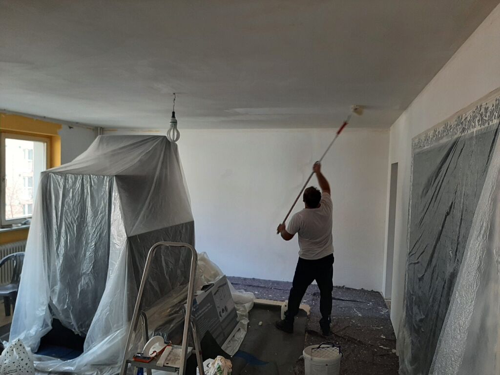 Handwerker streicht mit der Farbrolle die Decke eines Wohnraums, Möbel sind mit Folie abgedeckt